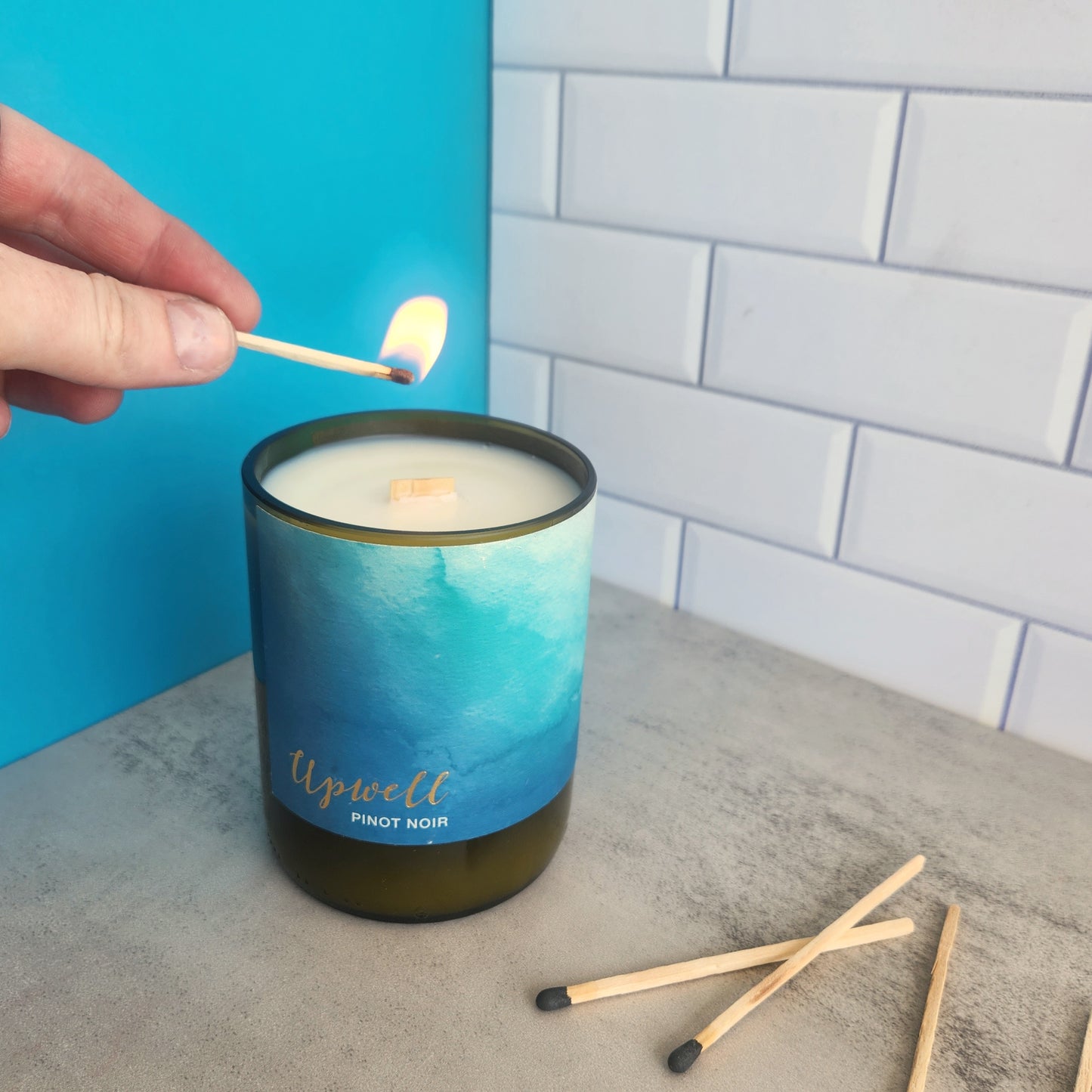White candle made in cut wine bottle. Label is blue watercolor and says Upwell Pinot Noir. Candle is being lit.