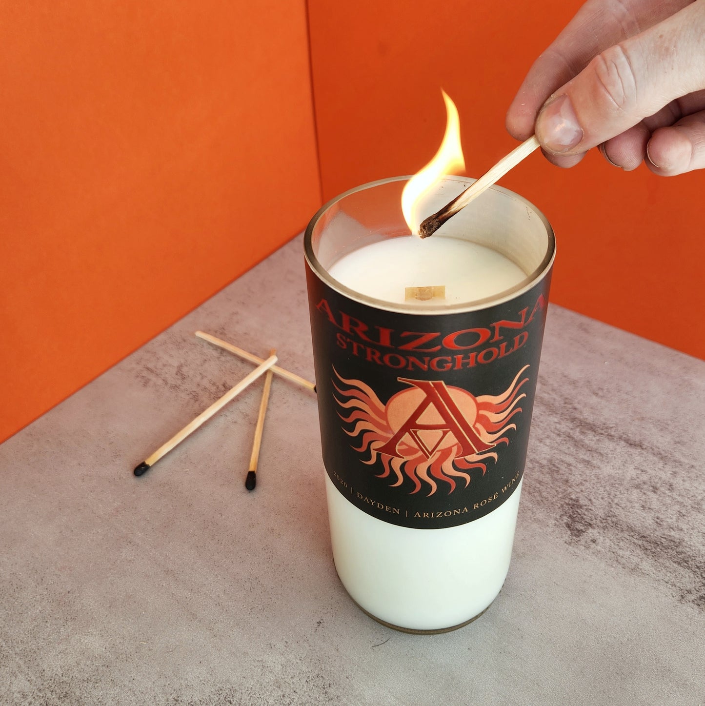 White candle with wooden wick in a cut off wine bottle. Label is black with an A surrounded by a stylized sun and states "Arizona Stronghold, 2020 Dayden Arizona Rose' Wine". Candle is being lit.