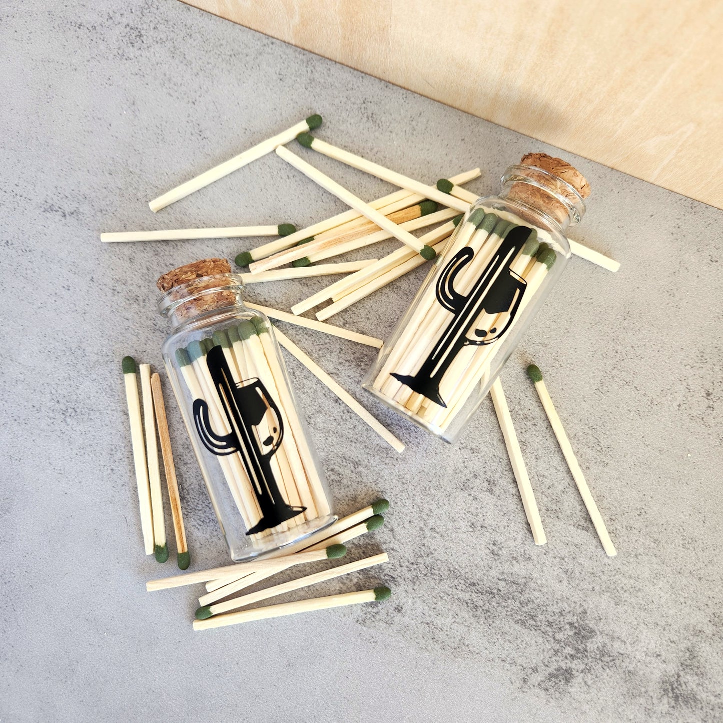 Overhead view of two glass bottles filled with 25 safety matches each, featuring the Cactus Vine logo and cork tops, with loose matches scattered around them on a textured surface.