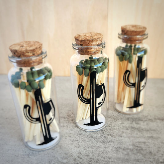 Three glass bottles filled with safety matches, each with a cork top and Cactus Vine logo, neatly arranged in a row on a textured background, showcasing their stylish design.