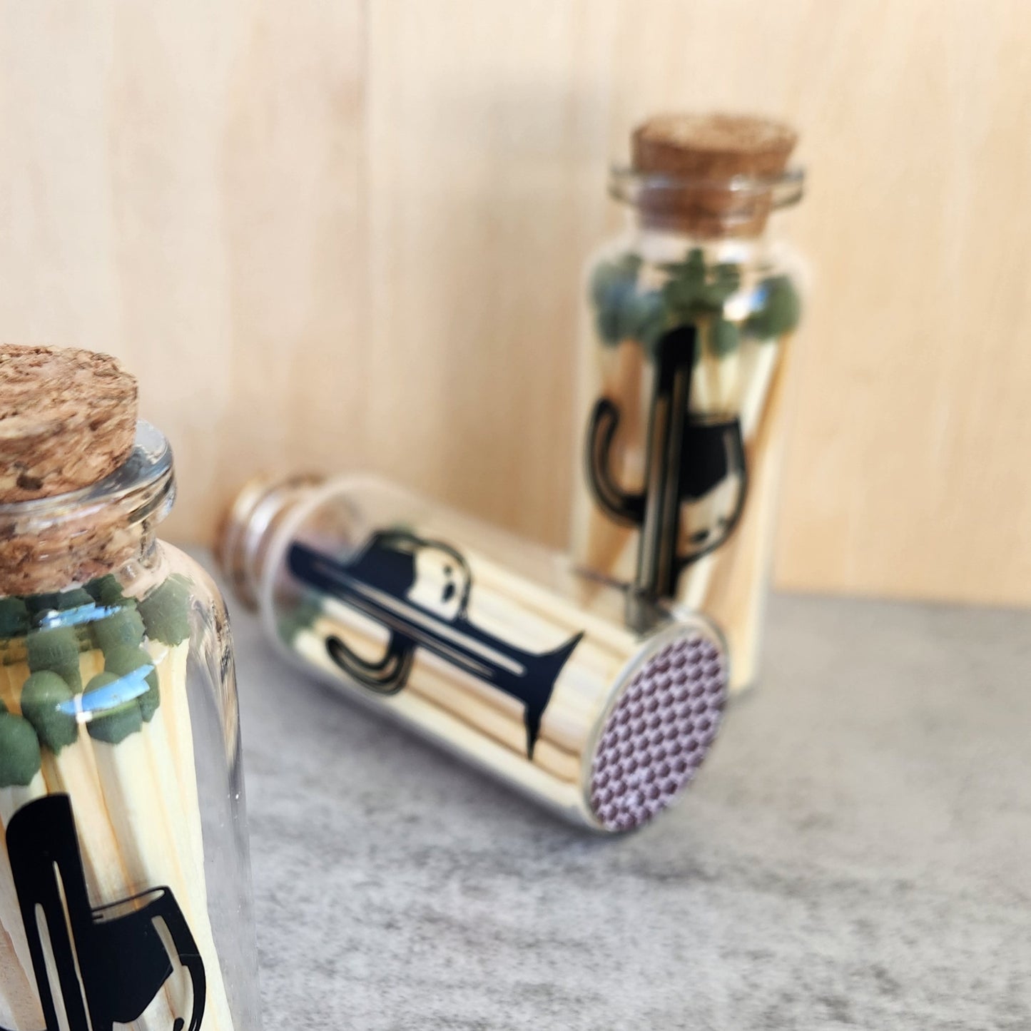 Close-up of glass bottles containing safety matches, highlighting the built-in striker pad on the bottom of one bottle and the unique Cactus Vine logo design on the front.