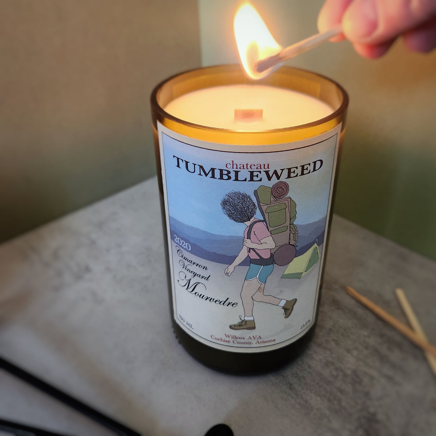 White candle made in a cut wine bottle. Label says Chateau Tumbleweed and has a drawing of a person walking through the desert wearing a backpack, with a tumbleweed for a head. There is a tent is the background. Candle is being lit.
