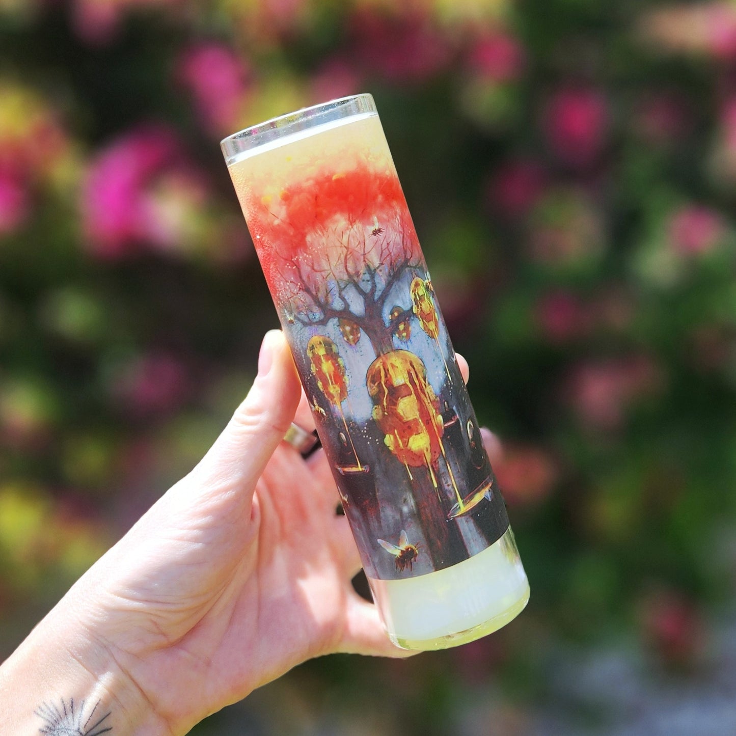 A tall, recycled glass candle with a vibrant artistic label featuring a surreal, autumnal scene of beehives among leafless trees under a fiery red and orange sky. A hand holds the candle against a blurred outdoor background of greenery and blue sky, emphasizing the detailed artwork and giving a seasonal, mystical feel.