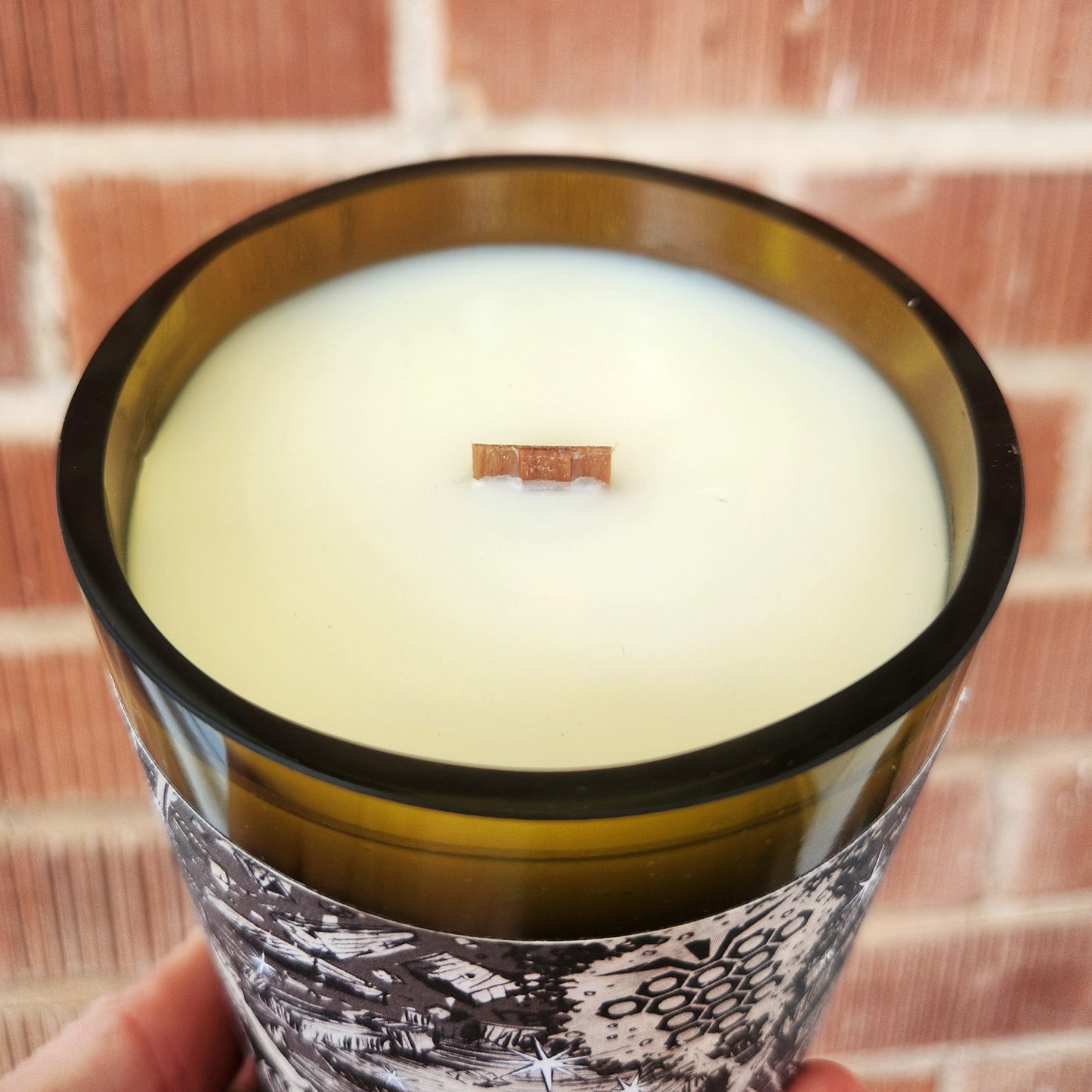 Close-up of the Rune wine bottle candle’s smooth rim and wooden wick, highlighting the creamy coconut soy wax and recycled glass vessel. This non-toxic candle catches the light in front of a red brick wall.
