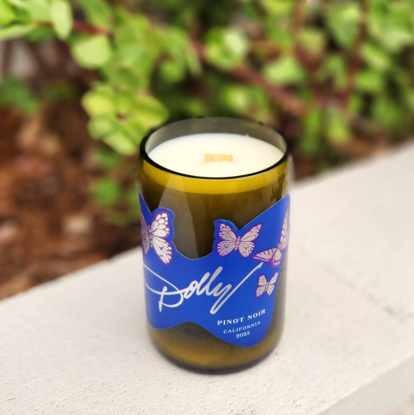 Angled top view of a hand-poured coconut soy candle housed in a recycled wine bottle with a vivid purple butterfly label and wooden wick centered in the creamy wax.