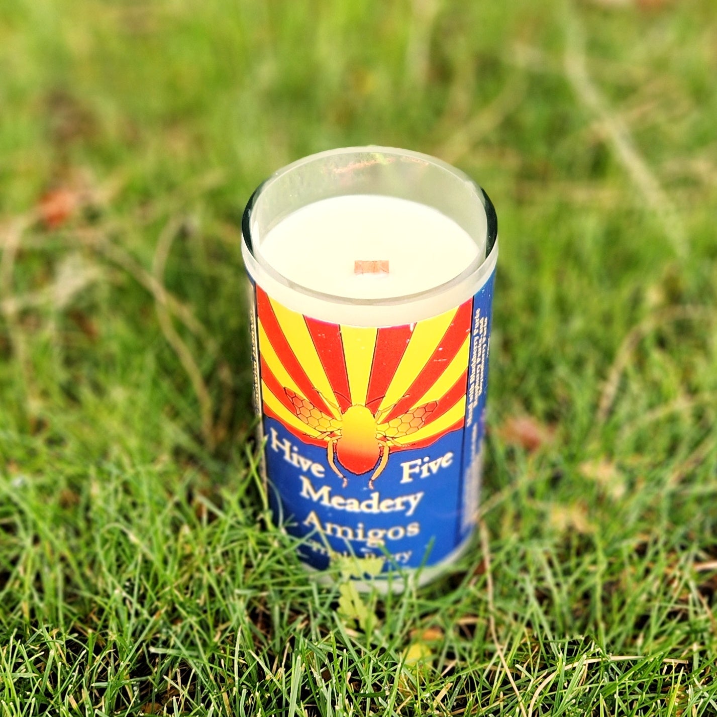 Angled view of a recycled mead bottle candle with a bright Arizona flag label and wooden wick, placed in lush outdoor grass.