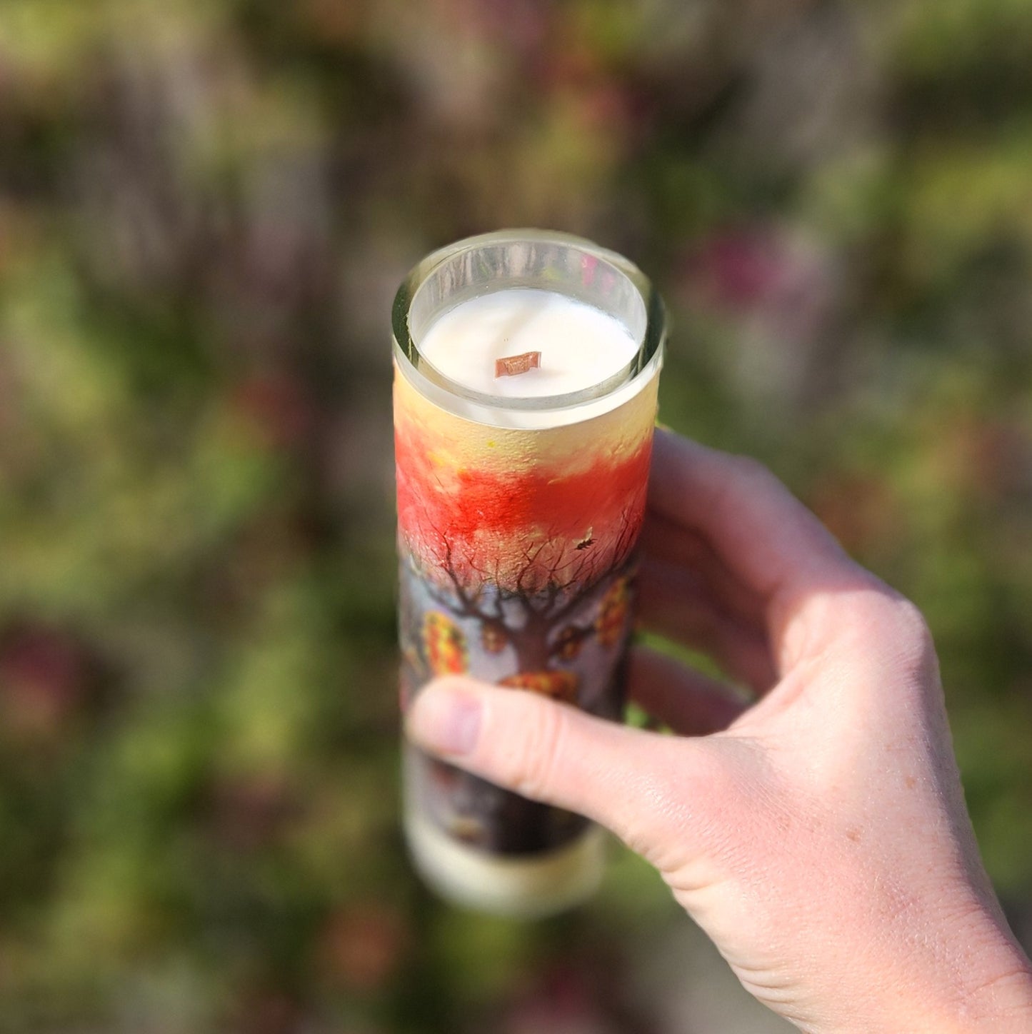 Angled close-up of the Rune Mead candle being held outdoors, showcasing the smooth coconut soy wax surface and wooden wick. The recycled bottle vessel features vivid sunset hues and autumn trees, emphasizing its hand-poured, non-toxic design.