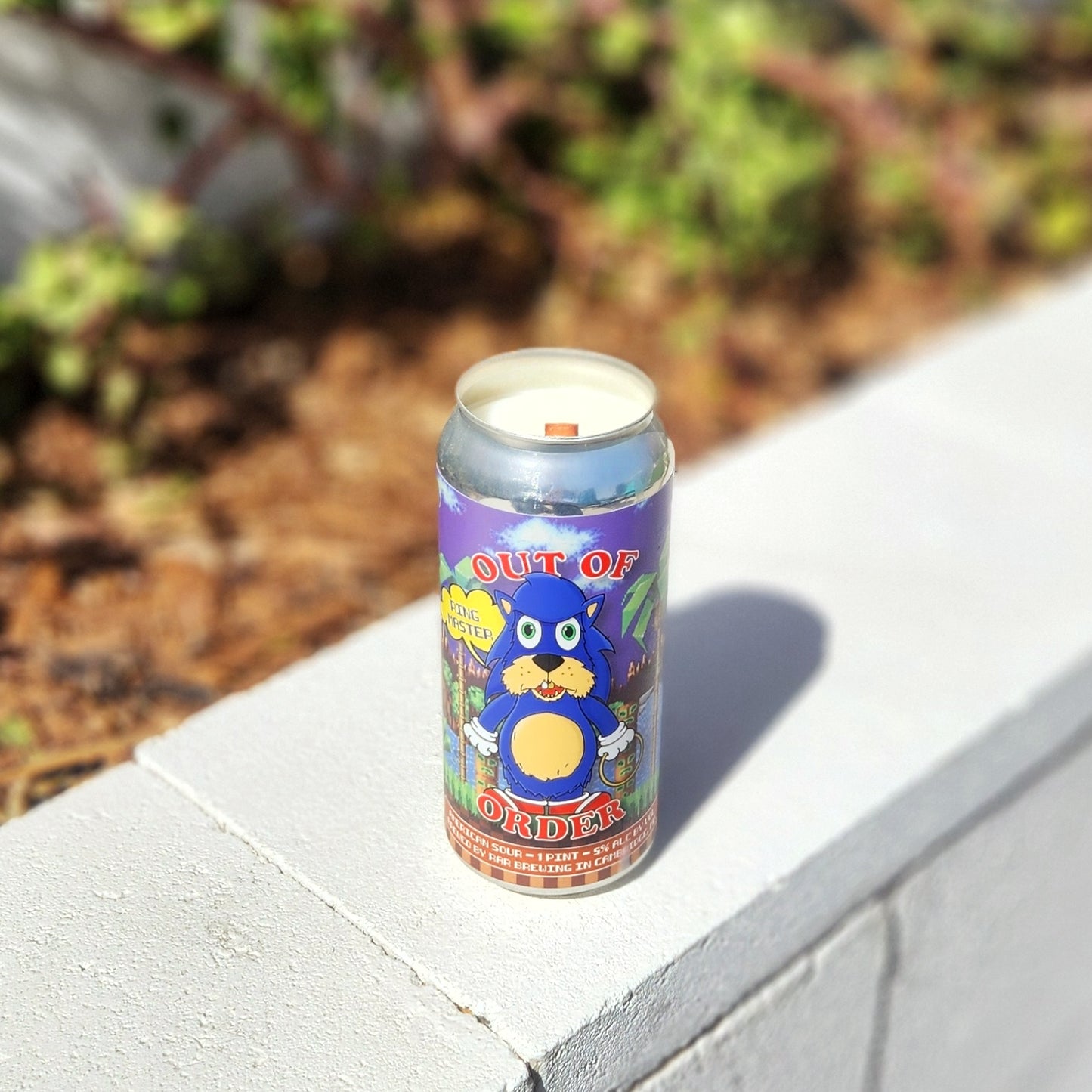 Angled view of a clean-burning coconut soy candle with a wooden wick, poured into a retro-themed recycled beer can. The colorful label features a bold blue cartoon character, offering a quirky and sustainable twist on traditional decor.
