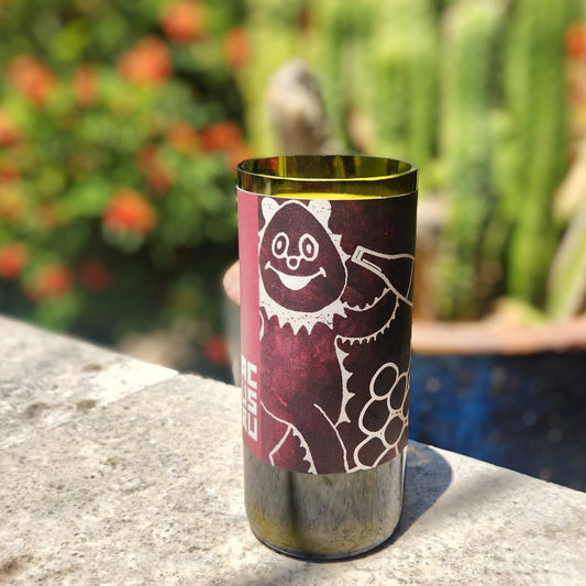 Eco-friendly, hand-poured coconut soy candle in a recycled wine bottle, featuring a whimsical white grape character on a dark red label; photographed outdoors with blurred greenery in the background.