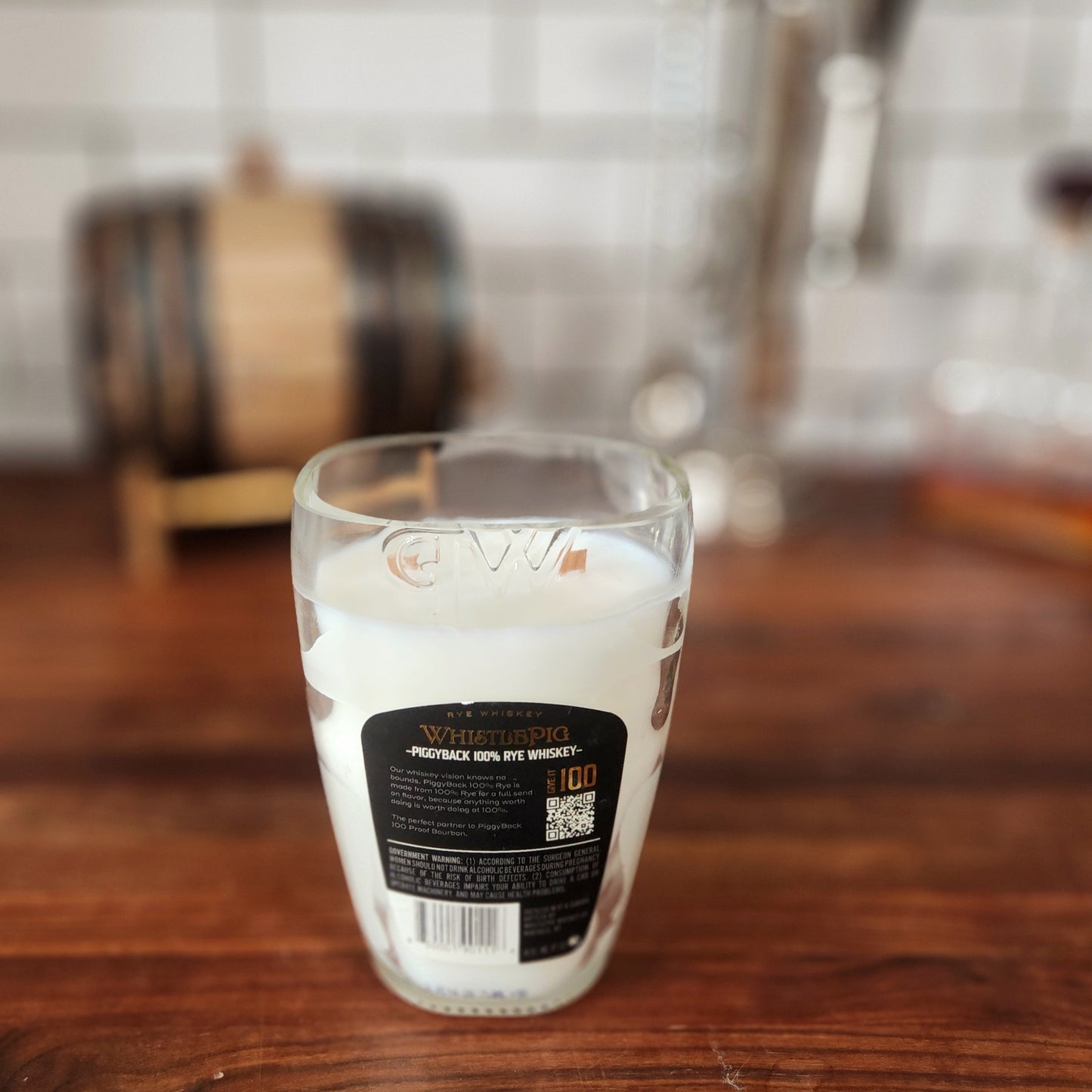 A hand-poured candle made with coconut soy wax and dual wooden wicks, housed in a recycled WhistlePig PiggyBack whiskey glass with its back label fully visible, placed on a rich wood counter with a mini whiskey barrel blurred in the background.