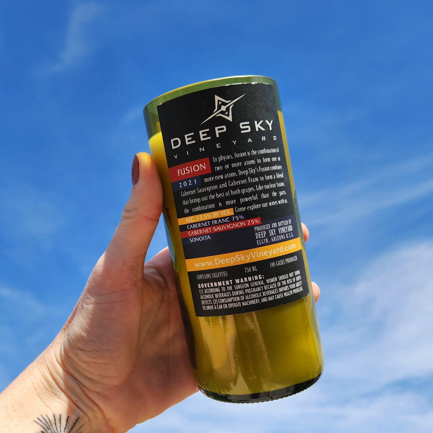 Back label view of the Fusion wine bottle candle from Deep Sky Vineyard, held up outdoors under a bright blue sky. The recycled bottle features a bold black label with red, white, and orange accents, emphasizing its clean-burning, handcrafted nature.
