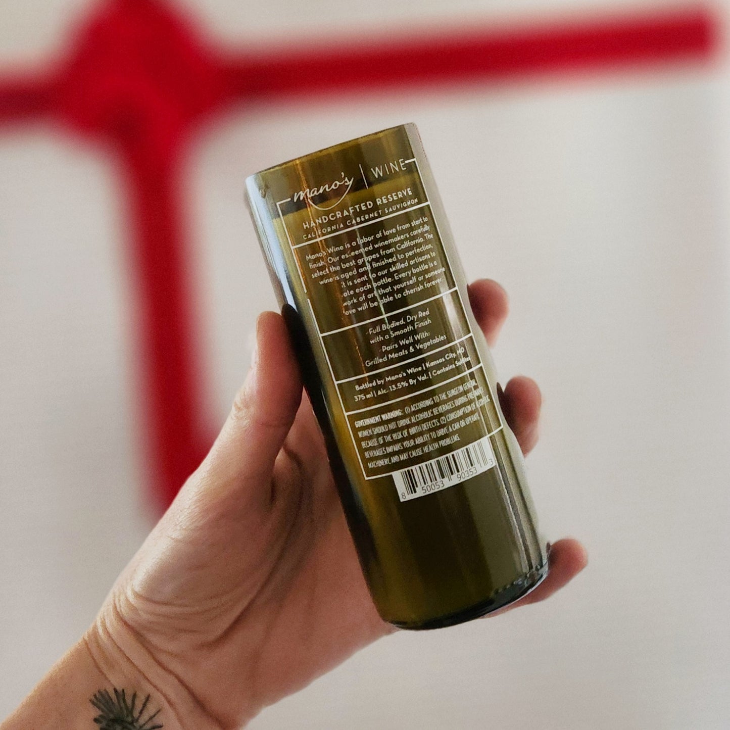 Back view of a recycled Mano’s Wine bottle candle featuring the original handcrafted reserve label, now filled with coconut soy wax and a wooden wick for a festive, eco-friendly holiday candle.