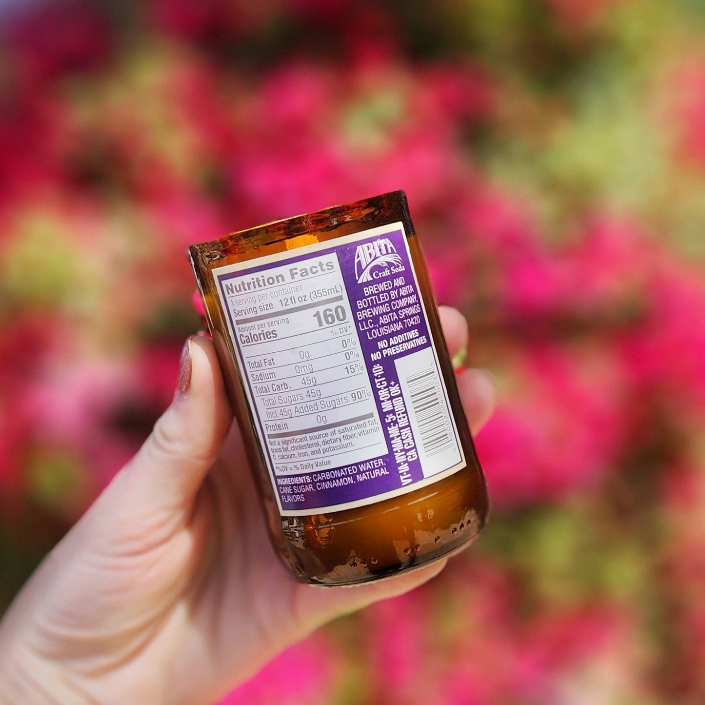 A hand holding a repurposed amber soda bottle candle in front of vibrant pink blooms. The label reveals the original nutritional facts from the soda brand. The festive coconut soy candle inside is hand poured with a wooden wick and housed in a smooth, upcycled glass vessel.
