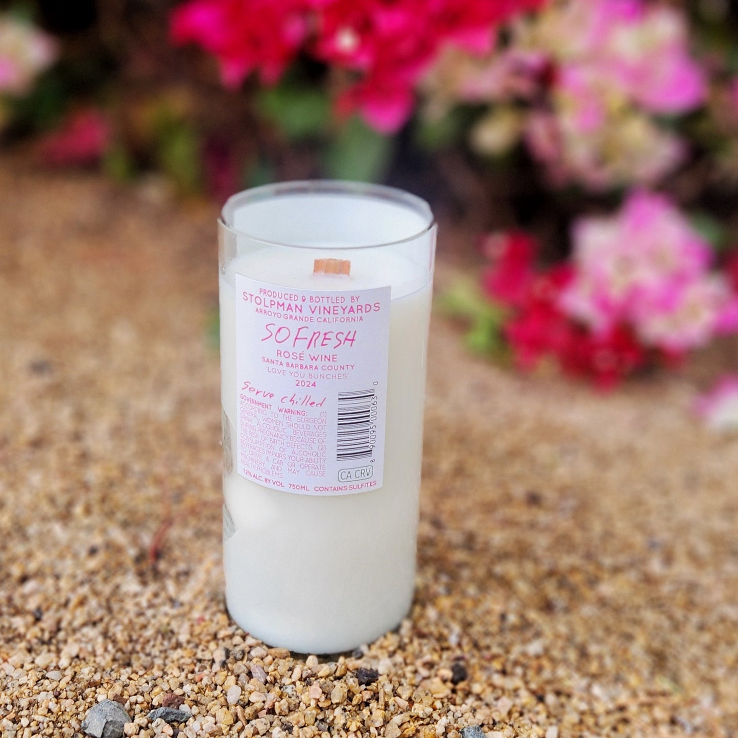 A clean-burning coconut soy wax candle housed in a recycled rosé wine bottle from Stolpman Vineyards, featuring a pink label that reads “So Fresh.” The eco-friendly candle is topped with a wooden wick and surrounded by vibrant pink flowers in the background.