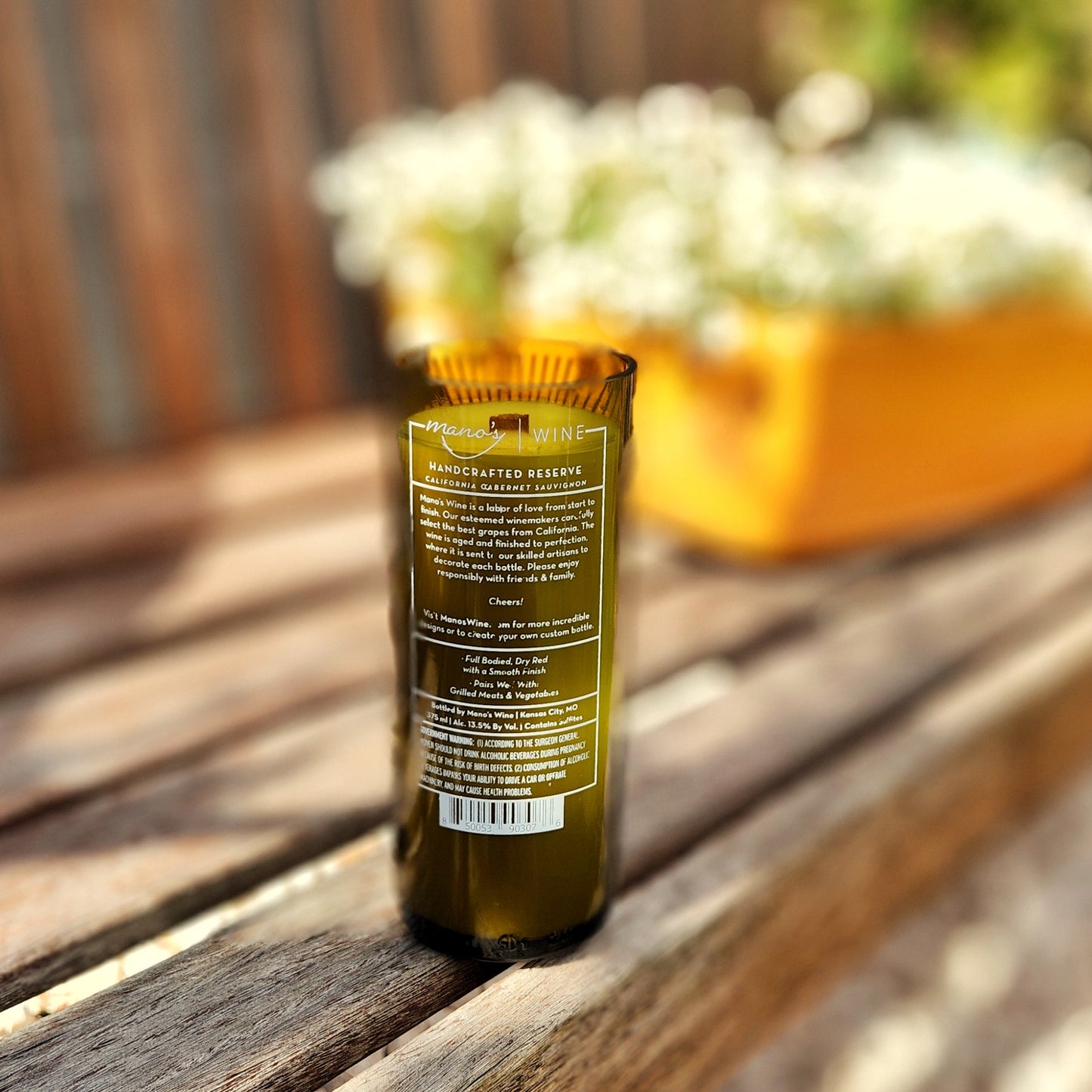 Back view of an upcycled wine bottle candle with visible wine label and eco-conscious branding. The recycled vessel is filled with clean-burning coconut soy wax and includes a wooden wick for a sustainable, non-toxic burn.