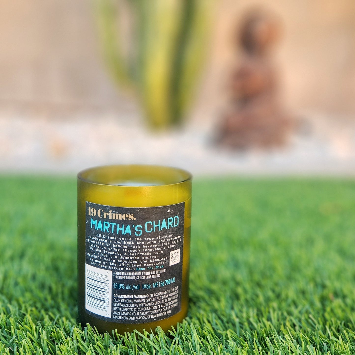 Back view of a clean-burning, eco-friendly wine bottle candle made from a recycled 19 Crimes bottle featuring the 'Martha’s Chard' label. The candle sits on green grass with blurred cacti in the background, emphasizing its sustainable and natural aesthetic.
