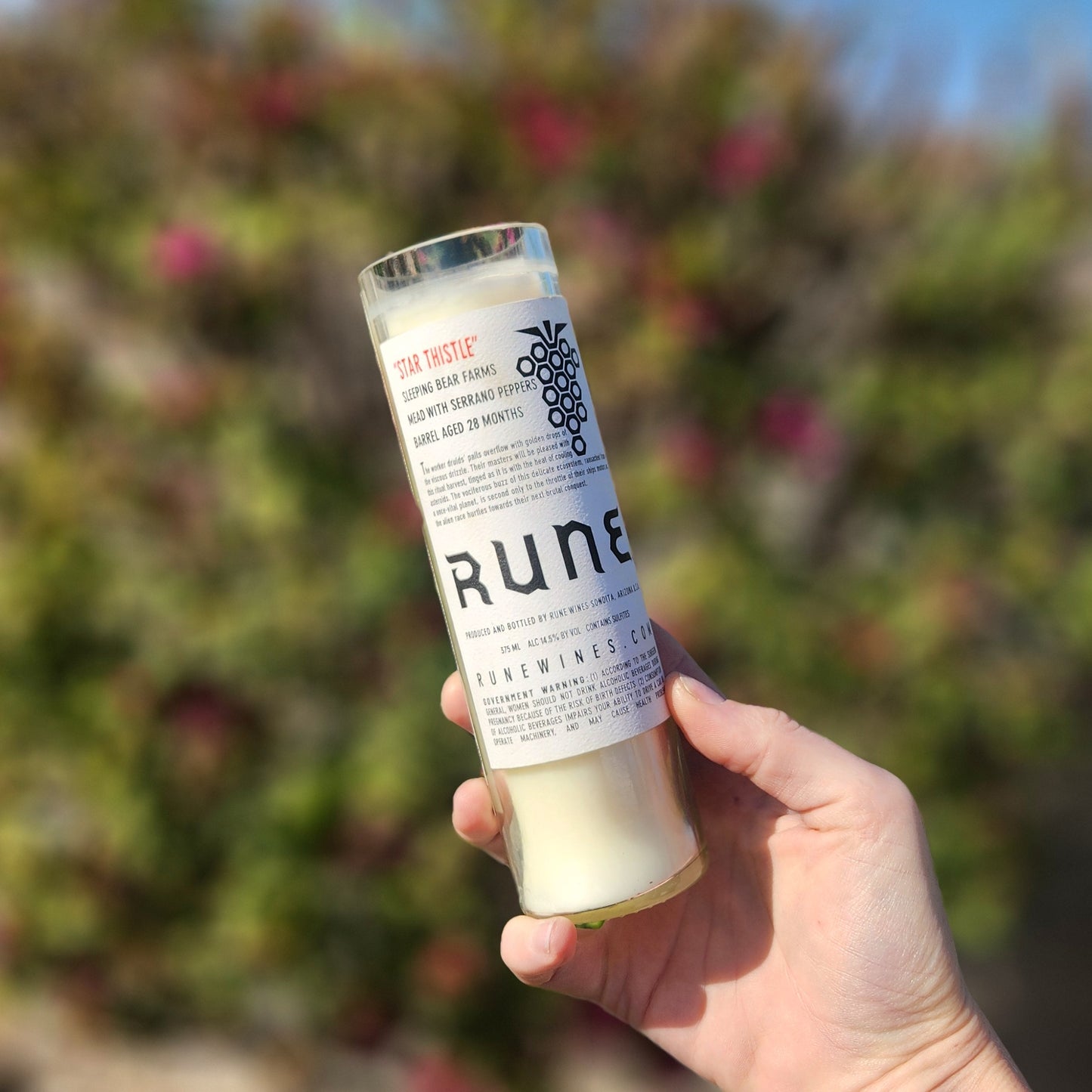 Back view of a tall Rune wine bottle candle labeled "Star Thistle" and "Mead with Serrano Peppers," held in hand with a floral garden backdrop. The label highlights the bottle’s origin and craft mead story, reinforcing the candle’s artisanal and eco-conscious appeal.