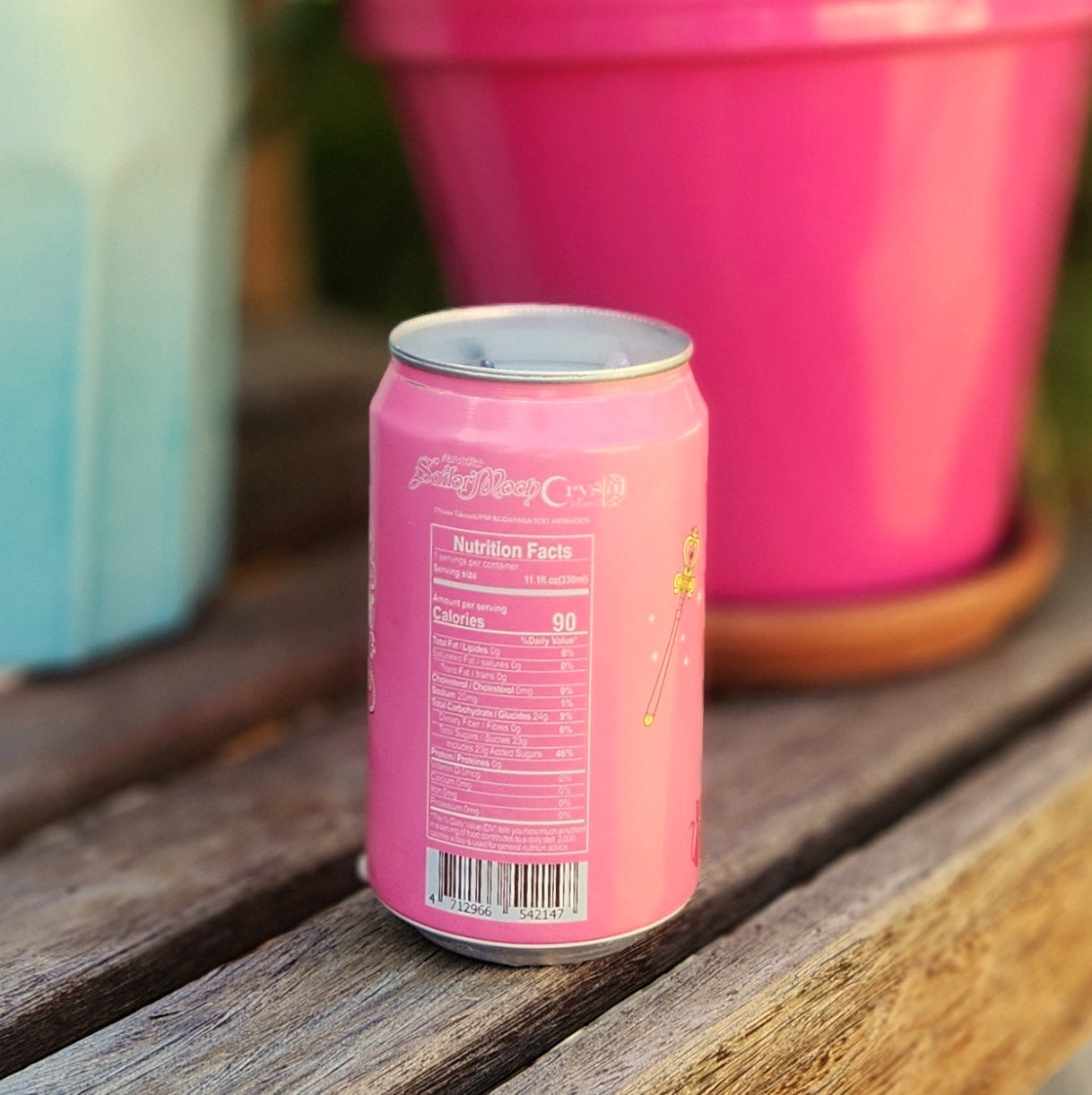 The back side of a pink Ocean Bomb can candle placed on a wooden bench, showing the nutrition facts and a small illustration of a magic wand, with a pink flower pot blurred in the background.