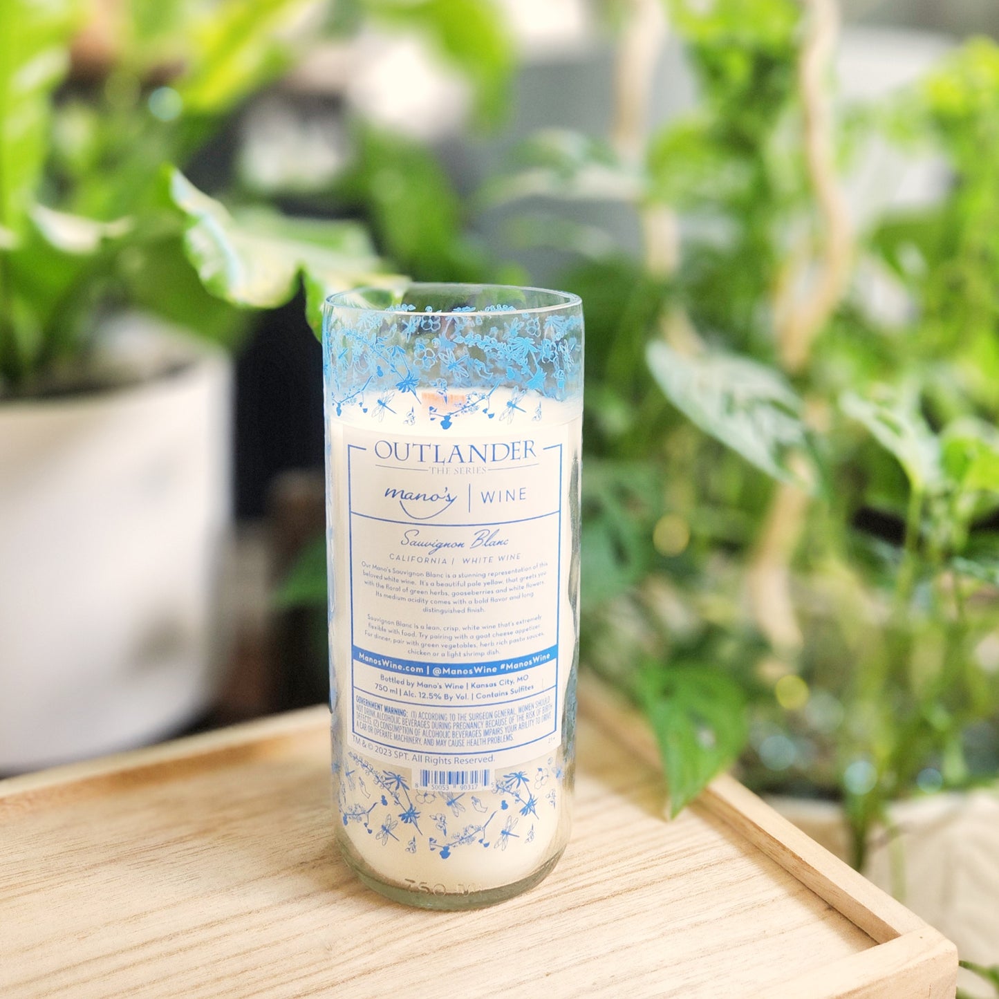 Hand-poured coconut soy wax candle in a recycled Outlander-themed wine bottle featuring delicate blue floral details and a soft white wax finish. This clean-burning, eco-friendly wine bottle candle is made with a wooden wick and natural ingredients, reflecting Cactus Vine’s signature sustainable style.