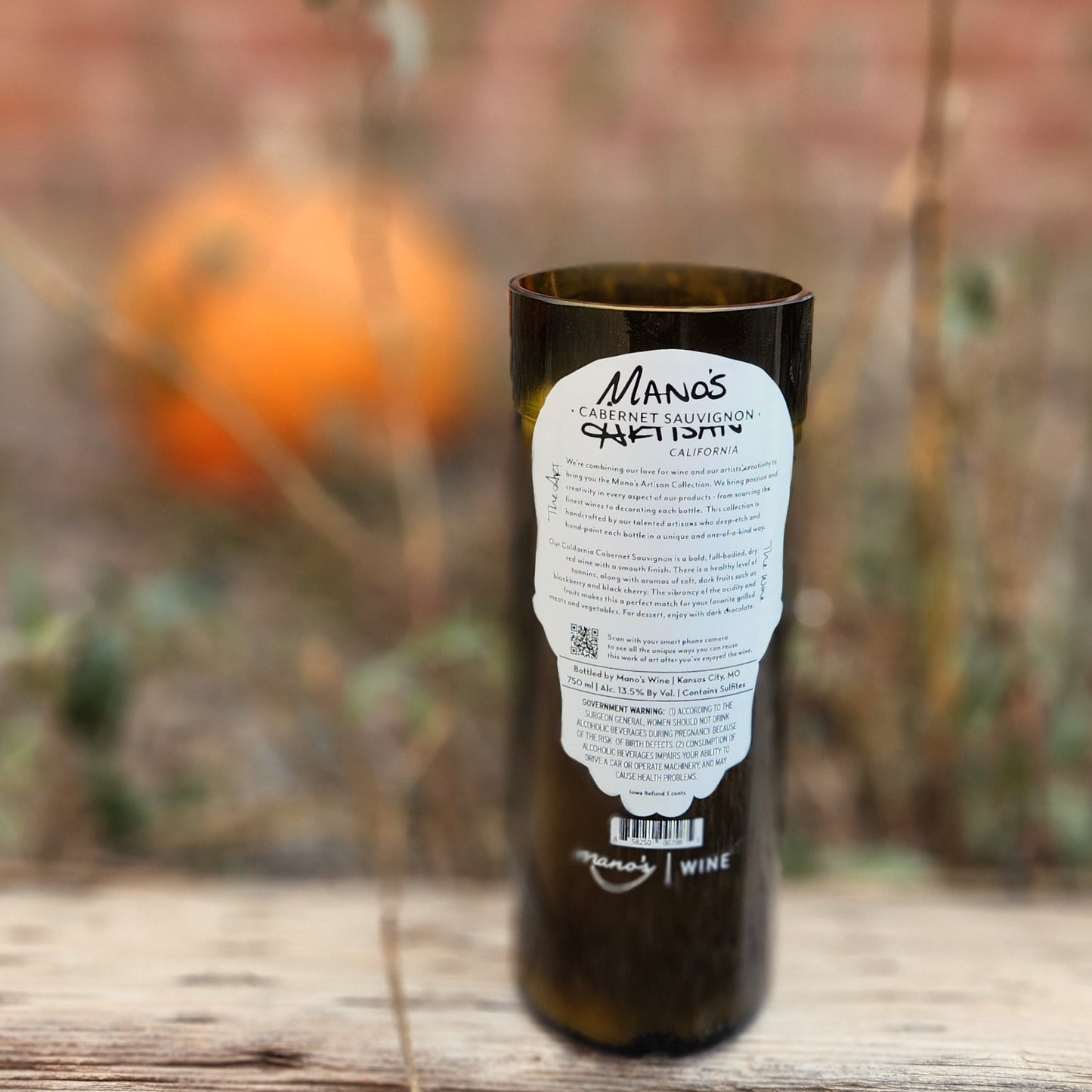 Back label of an eco-friendly wine bottle candle handcrafted from a Manos wine bottle, featuring vineyard info and set against an autumnal background with a pumpkin in soft focus.