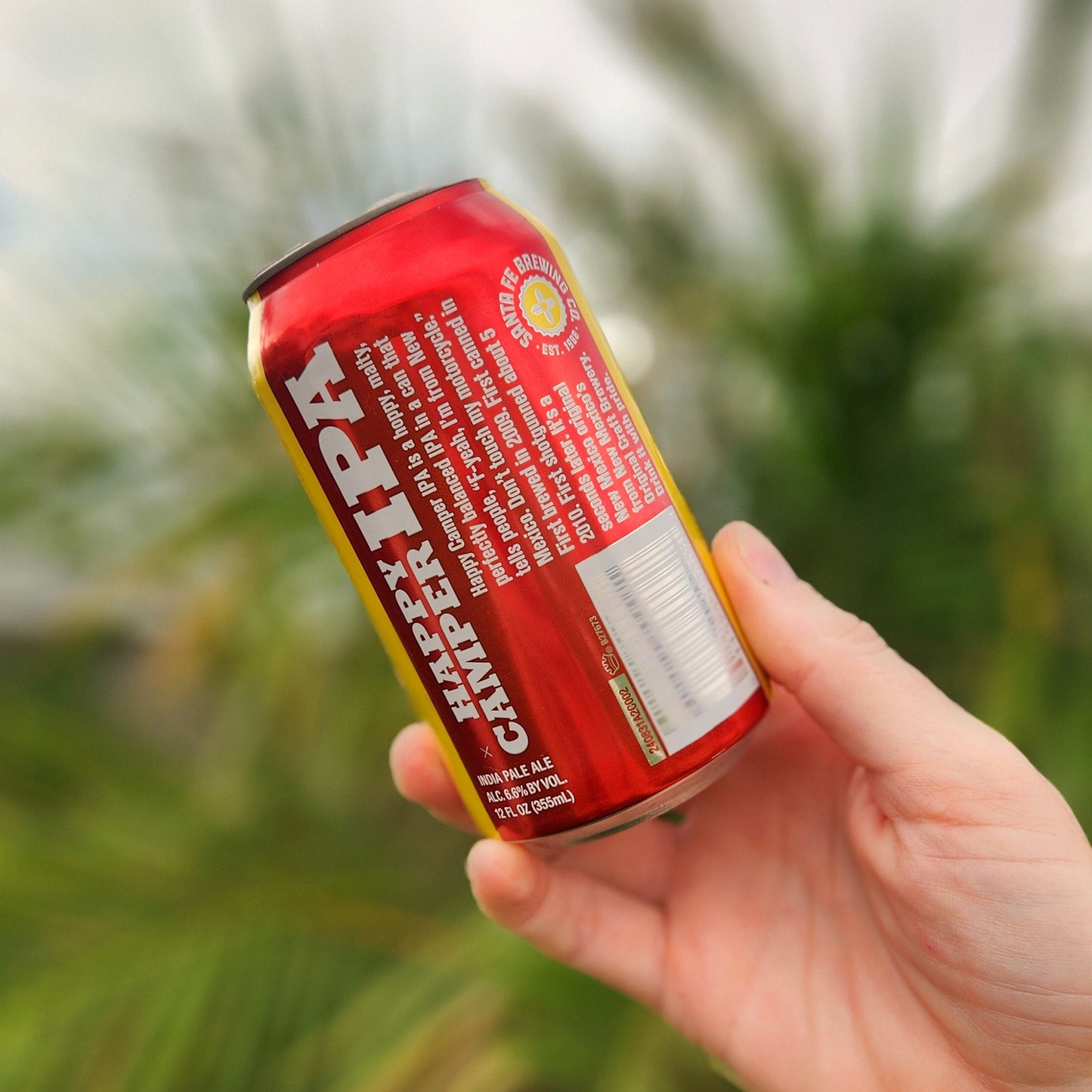 A hand holding a red and yellow Happy Camper IPA can from Santa Fe Brewing Co., with the back label and ingredients clearly visible. The background features blurred palm fronds and a cloudy sky, emphasizing the recycled nature of the can used for an upcycled candle.