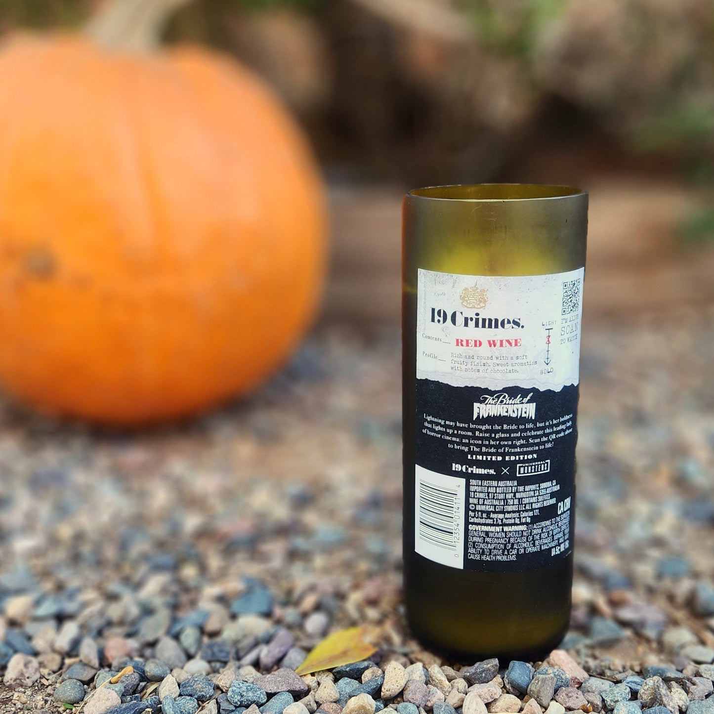 Back view of an eco-friendly wine bottle candle made from a recycled 19 Crimes red wine bottle. The label features themed artwork for “The Bride of Frankenstein,” blending gothic flair with a moody palette. Set outdoors on pebbled ground with a pumpkin blurred in the background, the shot highlights the smooth glass rim and sustainable craftsmanship of the upcycled vessel.