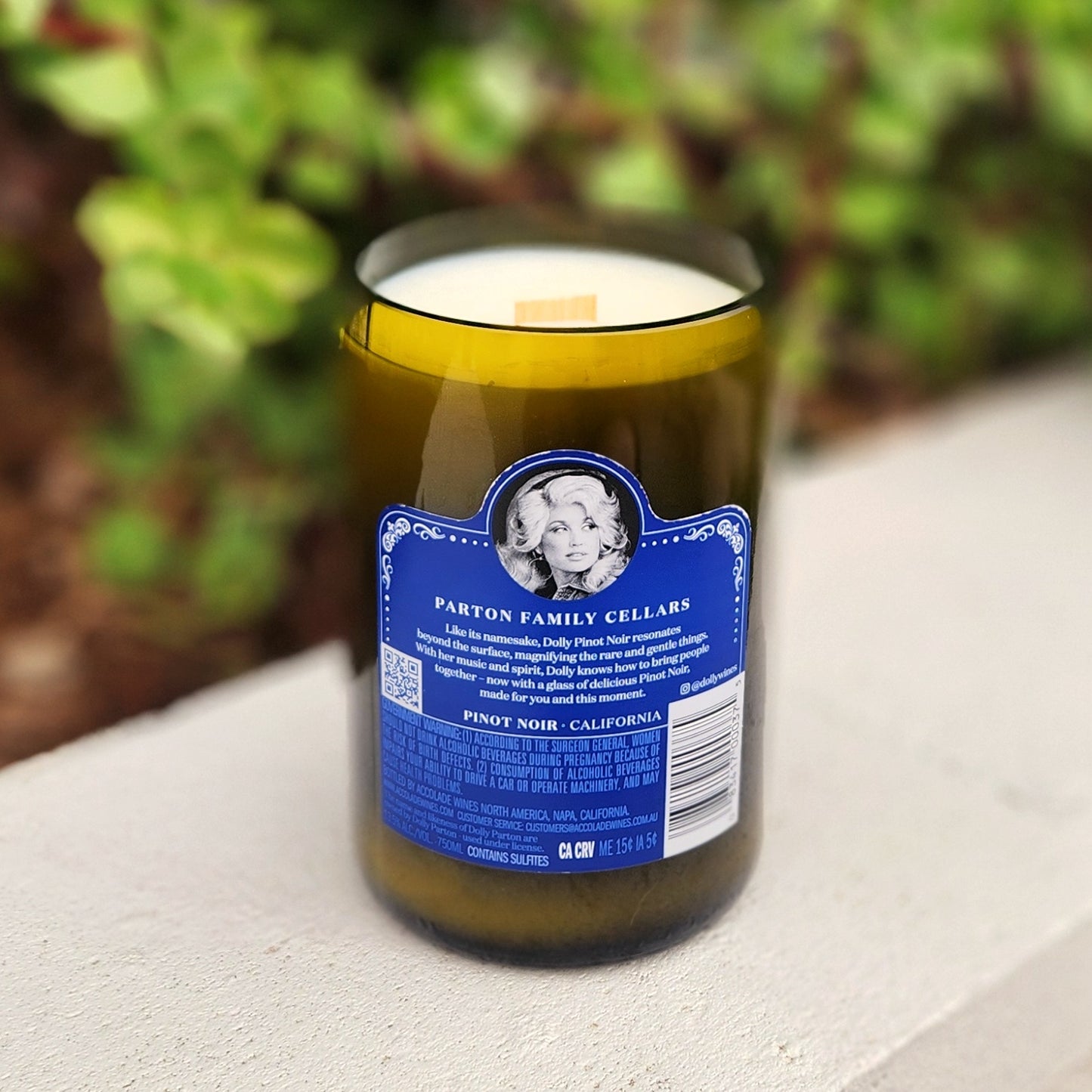 A recycled wine bottle candle featuring the blue back label of Parton Family Cellars with a black-and-white portrait of Dolly Parton and product description text, highlighting the eco-friendly upcycled glass and clean-burning coconut soy wax inside.