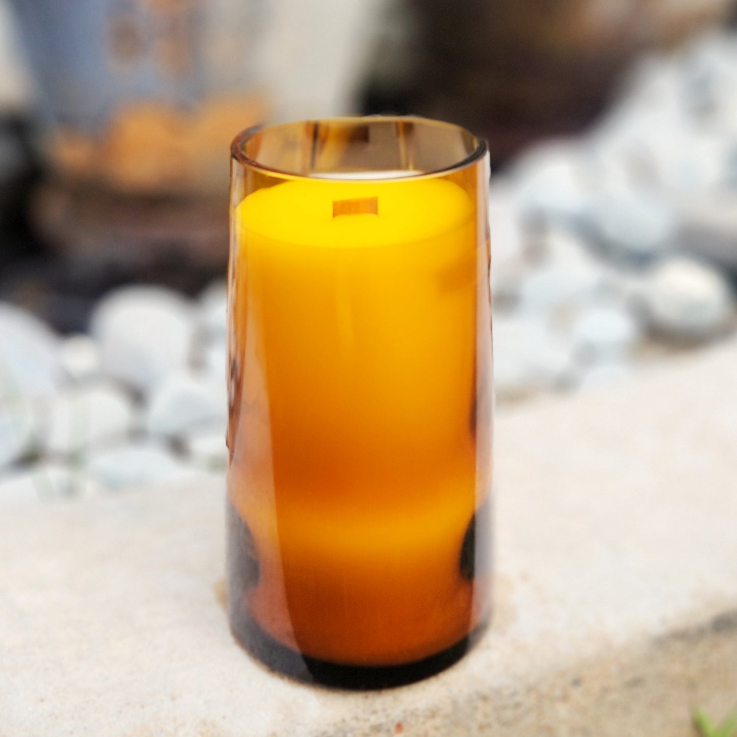 A clean-burning, coconut soy wax candle with a wooden wick, hand-poured into an amber recycled bottle, shown upright on a smooth stone surface.