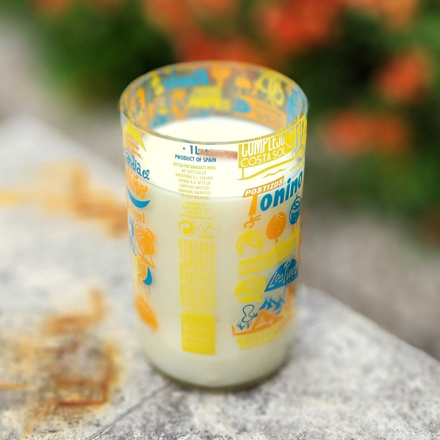 A hand-poured coconut soy wax candle in a recycled wine bottle with a vibrant retro-style travel label in yellow, orange, and blue, featuring whimsical graphics and playful fonts; the candle is placed outdoors on a sunlit stone surface.