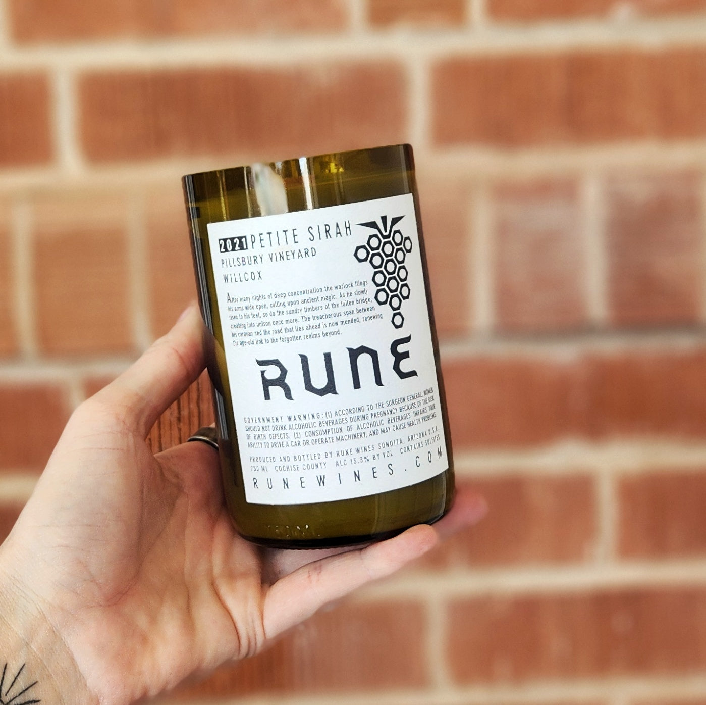 Eco-friendly candle in a recycled wine bottle featuring the Rune Petite Sirah label, hand-poured with clean-burning coconut soy wax being held in front of a red brick wall.