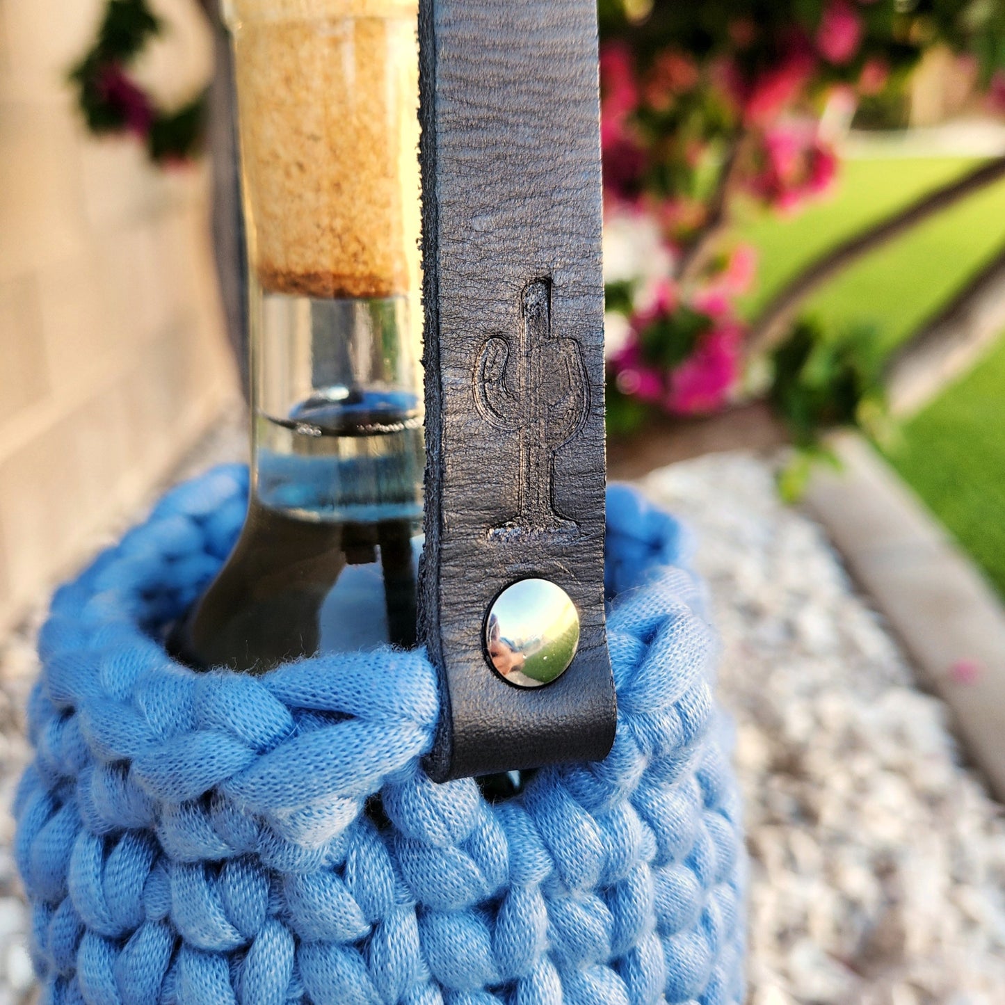 A close-up of a wine bottle nestled in a denim blue crocheted carrier with a black leather strap, featuring a stamped cactus design. The cactus branding on the strap pops against the soft yarn texture, highlighting the handcrafted details and rustic elegance of the wine bottle holder.