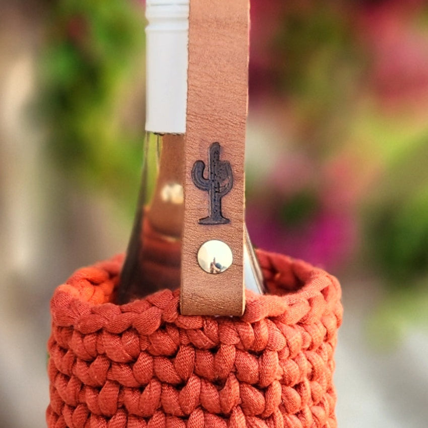 A close-up of a burnt orange crochet wine bottle carrier with a tan leather handle, featuring a branded cactus wine logo and silver rivet. The bottle inside has a white cap, and the background is softly blurred with greenery and pink flowers, adding a vibrant springtime touch.