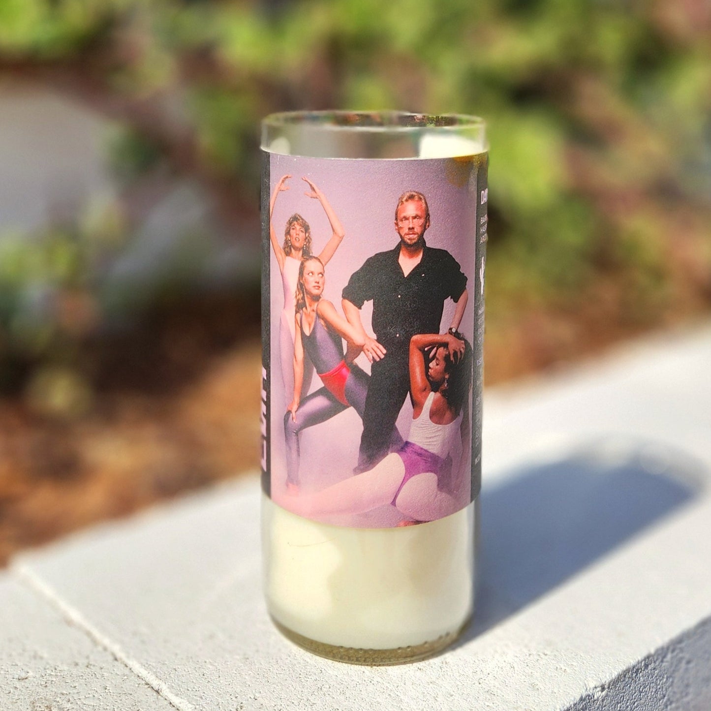 A hand-poured coconut soy wine bottle candle crafted from a recycled Arizona wine bottle. The label features a retro dance-inspired design with a dramatic group pose, adding a bold and nostalgic aesthetic. This eco-friendly wooden wick candle has a polished glass rim, highlighting its artisan craftsmanship. Placed outdoors on a sunlit concrete ledge with a blurred green backdrop, this sustainable wine bottle candle is a perfect blend of artistry, nostalgia, and sustainability.