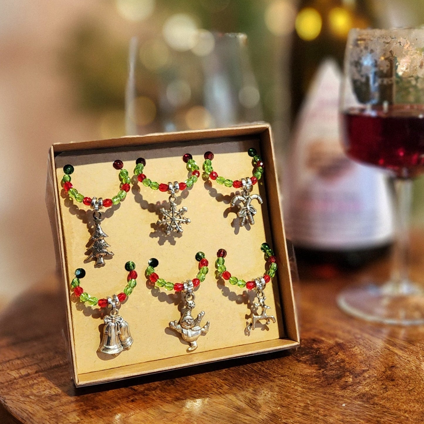 Festive wine glass charms featuring colorful beads and holiday-themed pendants, including a Christmas tree, snowflake, and bells, displayed in a gift box with a wine glass and bottle in the background.
