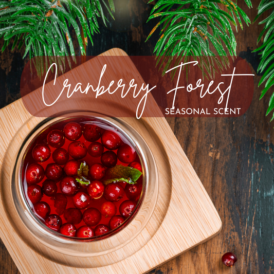 A cozy flat-lay featuring a bowl of fresh cranberries in rich red liquid, garnished with mint, placed on a wooden coaster over a rustic wood surface. Evergreen branches frame the top edge of the image, adding a festive, forest-like feel. A translucent maroon overlay displays the words "Cranberry Forest – Seasonal Scent" in elegant white script.