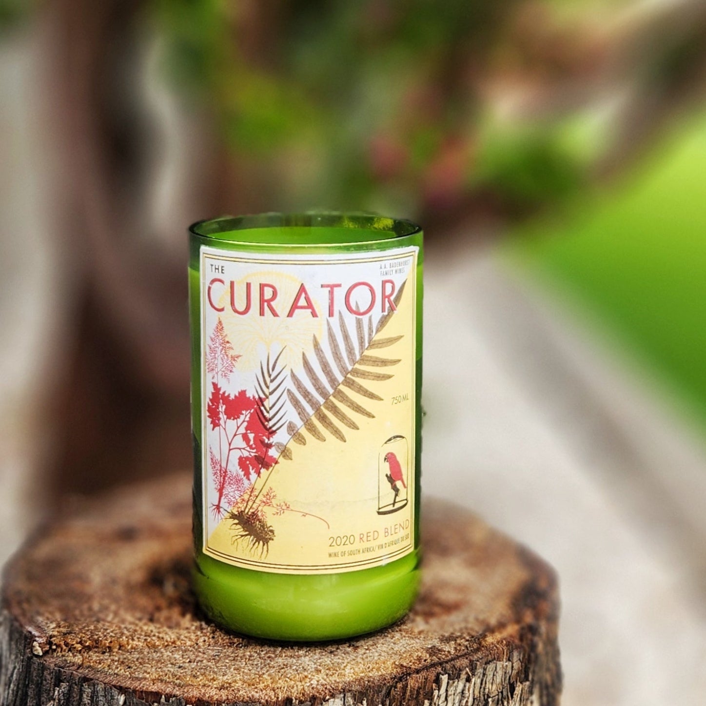 A hand-poured coconut soy wine bottle candle crafted from a recycled The Curator Red Blend 2020 bottle. The label features a nature-inspired design with delicate ferns and foliage in earthy tones, evoking a botanical aesthetic. This eco-friendly wooden wick candle has a smooth, polished glass rim, showcasing expert craftsmanship. Placed on a rustic tree stump with a blurred green garden in the background, this sustainable wine bottle candle is perfect for nature lovers, wine enthusiasts, and eco-conscious s