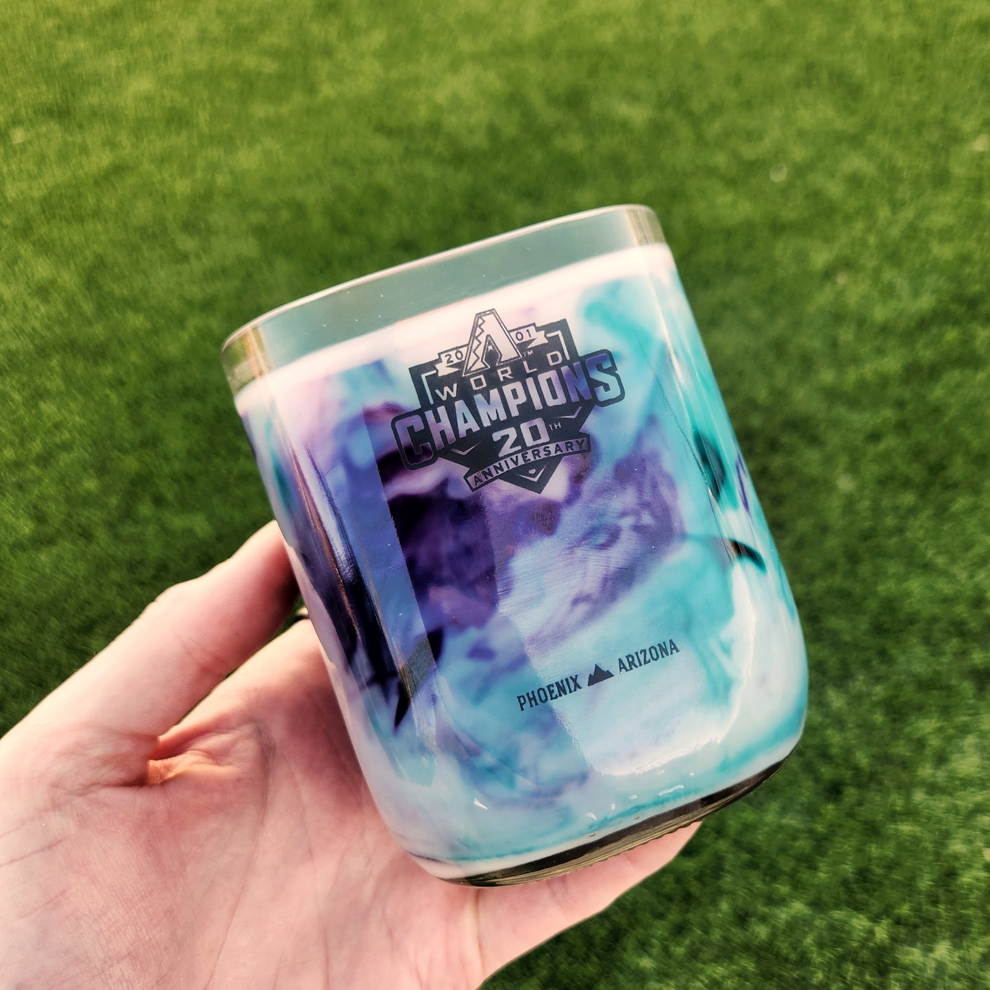 Close-up of a hand holding a clean-burning wine bottle candle featuring a AZ Diamondbacks 20th Anniversary Champions logo, repurposed into a vibrant coconut soy wax candle with a wooden wick.