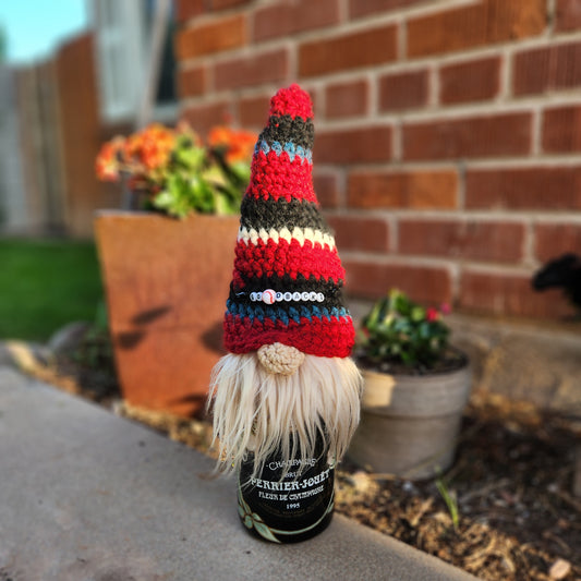 A crocheted wine bottle gnome in red, black, and white with a beaded “DBacks” charm on the hat. Sits atop a champagne bottle.