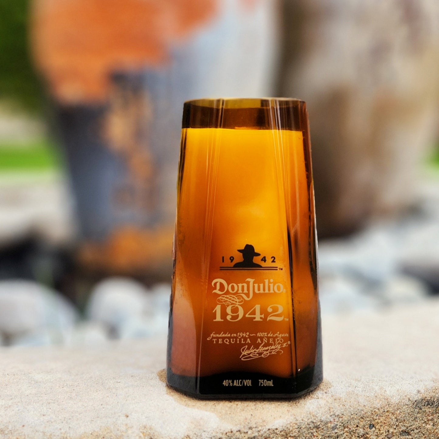 A hand-poured coconut soy candle housed in a recycled Don Julio 1942 tequila bottle, featuring its signature amber glass and gold lettering. This eco-friendly wooden wick candle has a smooth, polished rim, showcasing expert craftsmanship. Set outdoors on a stone surface with a blurred background of rustic pottery and greenery, this upcycled tequila bottle candle is a unique addition for tequila lovers and sustainable décor enthusiasts.