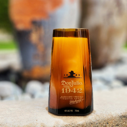 A hand-poured coconut soy candle housed in a recycled Don Julio 1942 tequila bottle, featuring its signature amber glass and gold lettering. This eco-friendly wooden wick candle has a smooth, polished rim, showcasing expert craftsmanship. Set outdoors on a stone surface with a blurred background of rustic pottery and greenery, this upcycled tequila bottle candle is a unique addition for tequila lovers and sustainable décor enthusiasts.