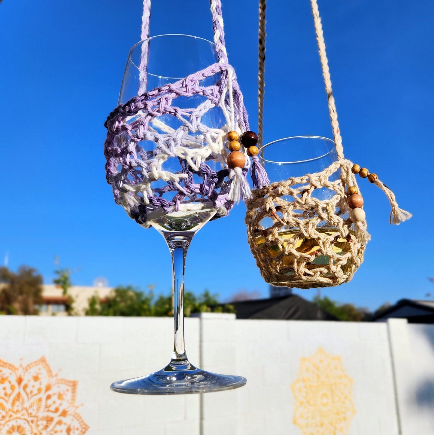 Two crochet wine glass lanyards hang side-by-side against a blue sky—one purple and white, the other tan. Both feature beaded details and are suspended in front of a white outdoor wall with soft orange mandala patterns.