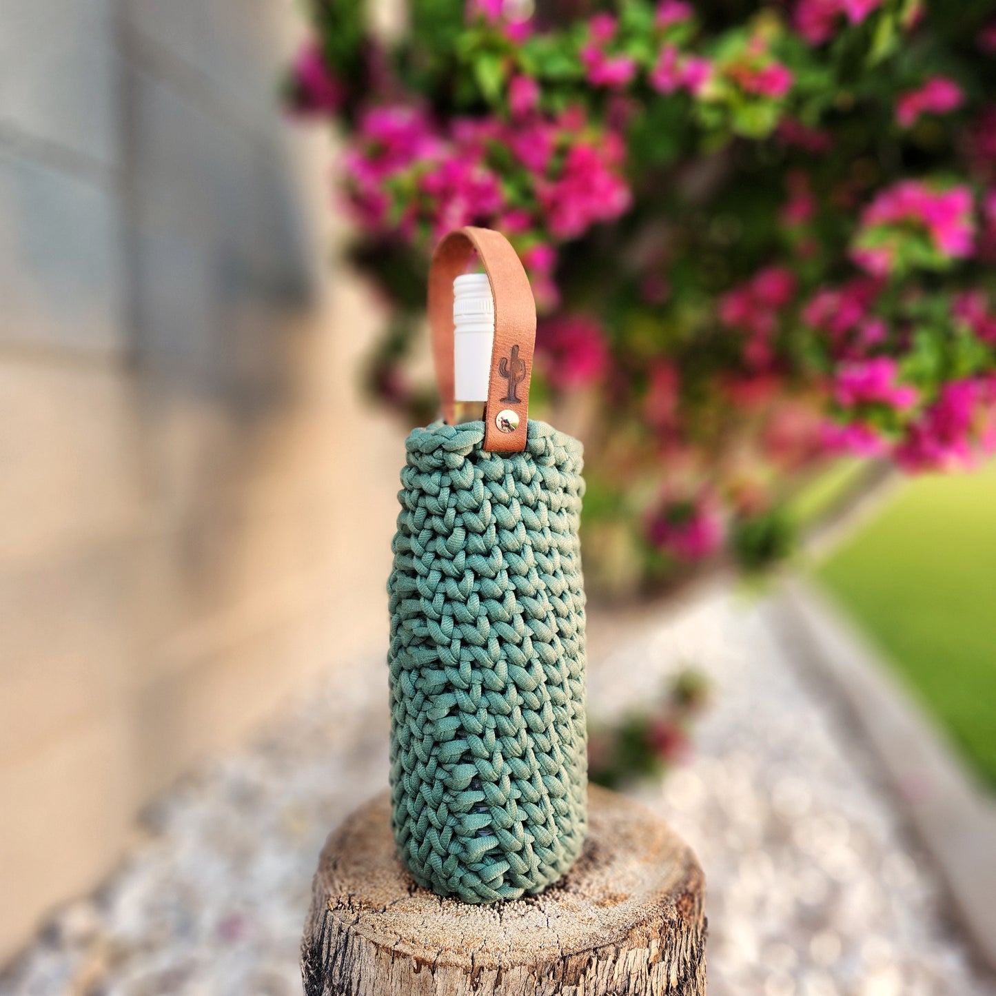 A hand-crocheted wine bottle carrier in a muted evergreen color, holding a white-capped bottle and featuring a tan leather handle stamped with a cactus design. The carrier rests on a tree stump outdoors, framed by soft-focus pink bougainvillea and garden greenery for a cozy, rustic vibe.