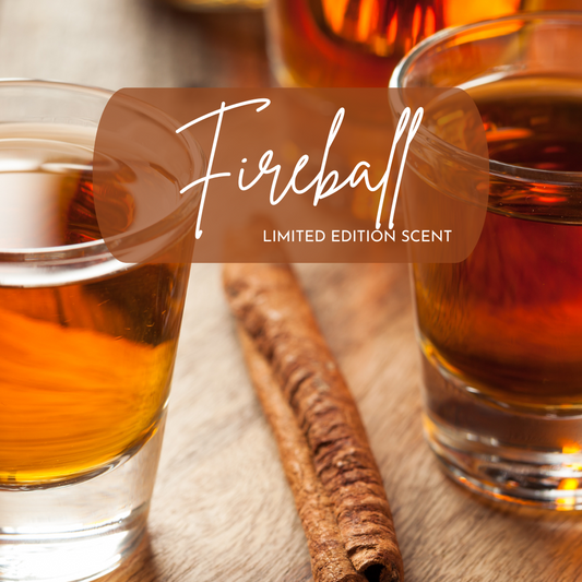 Close-up of two shot glasses filled with amber-colored whiskey beside a cinnamon stick on a wooden surface, with warm lighting highlighting the rich tones. Overlaid text reads “Fireball – Limited Edition Scent,” suggesting a spicy cinnamon and whiskey-inspired fragrance.