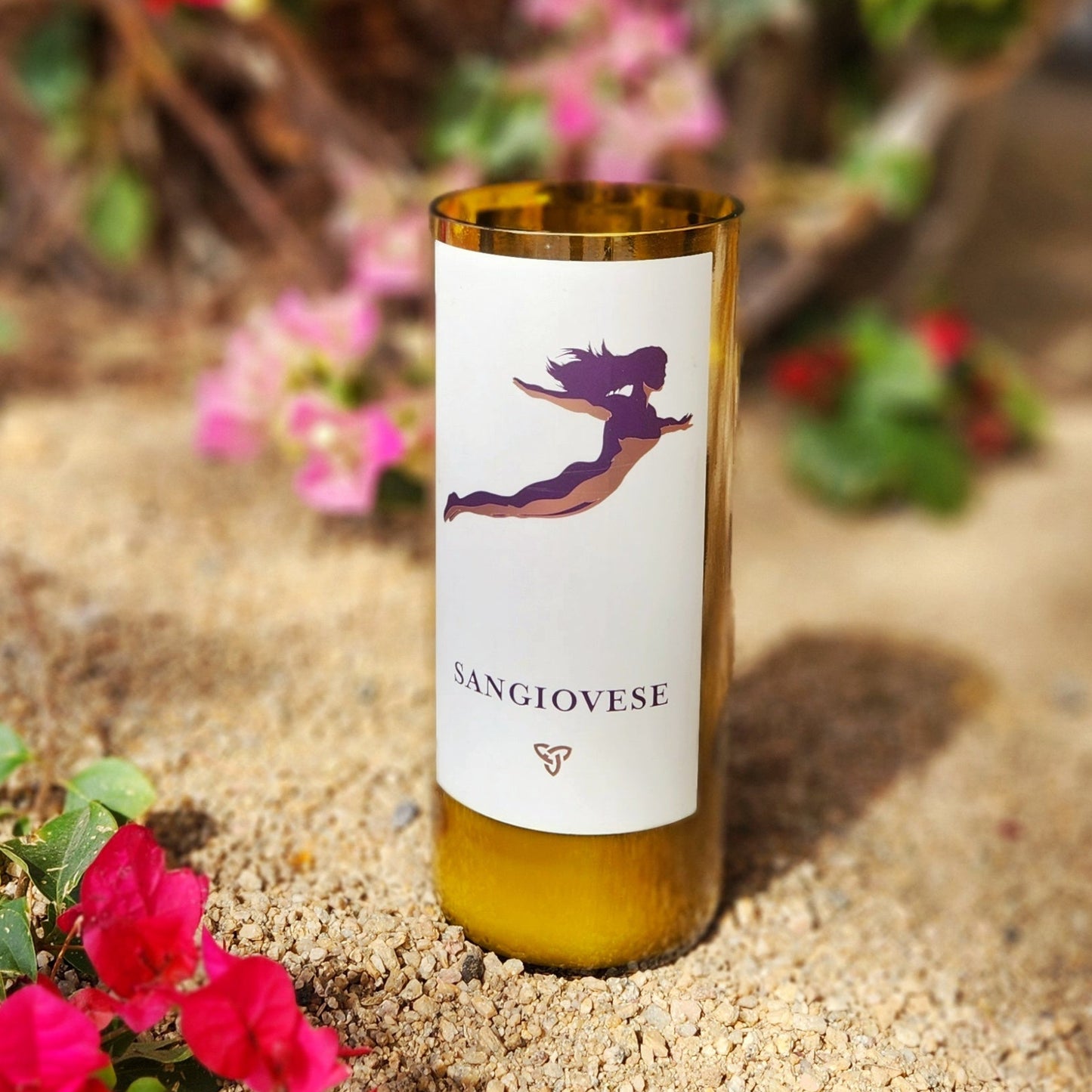 A hand-poured coconut soy wine bottle candle housed in a recycled Flying Leap Vineyards Sangiovese bottle. The label features an artistic silhouette of a woman in flight, adding a whimsical touch to this eco-friendly wooden wick candle. The smooth, polished glass rim highlights its artisan craftsmanship. Placed in a desert-inspired setting with vibrant pink bougainvillea flowers and sandy earth, this sustainable wine bottle candle beautifully blends nature, wine, and sustainability.