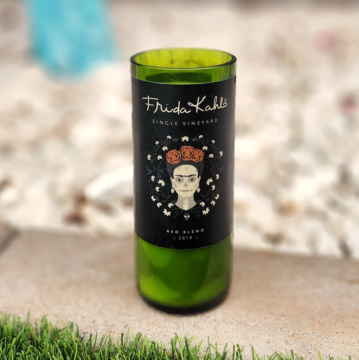 Non-toxic candle in a recycled wine bottle featuring Frida Kahlo’s portrait with floral crown and white flowers on a matte black label, filled with coconut soy wax and topped with a crackling wooden wick.