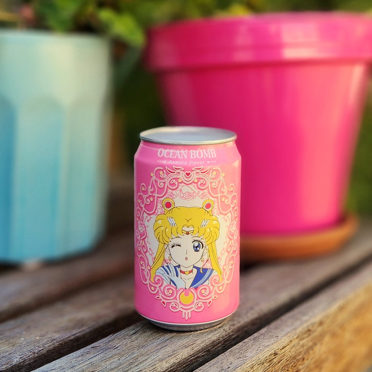 A pink Ocean Bomb can featuring a winking anime character with blonde hair and a cat atop her head, transformed into a recycled candle, shown outdoors on a wooden bench next to bright pink and blue planters.