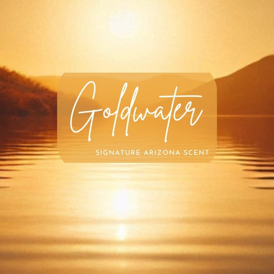 A warm, golden-hued sunset reflects over rippling lake water, framed by gentle desert hills in silhouette. The words “Goldwater – Signature Arizona Scent” are overlaid in elegant white script on a semi-transparent amber banner, evoking the scent's inspiration from Arizona’s golden lakes at dusk.