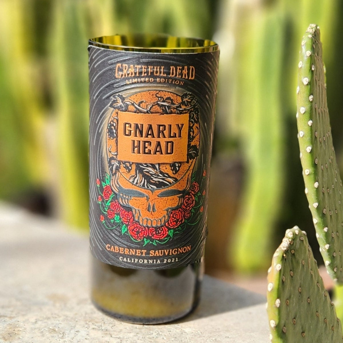 A hand-poured coconut soy wine bottle candle crafted from a recycled wine bottle. This Grateful Dead limited edition label features a bold skull design surrounded by roses and intricate artwork, evoking a rock-and-roll aesthetic. This eco-friendly wooden wick candle has a smooth, polished glass rim, highlighting expert craftsmanship. Placed on a sunlit stone surface with a cactus-filled desert background, this sustainable wine bottle candle is perfect for Grateful Dead fans.