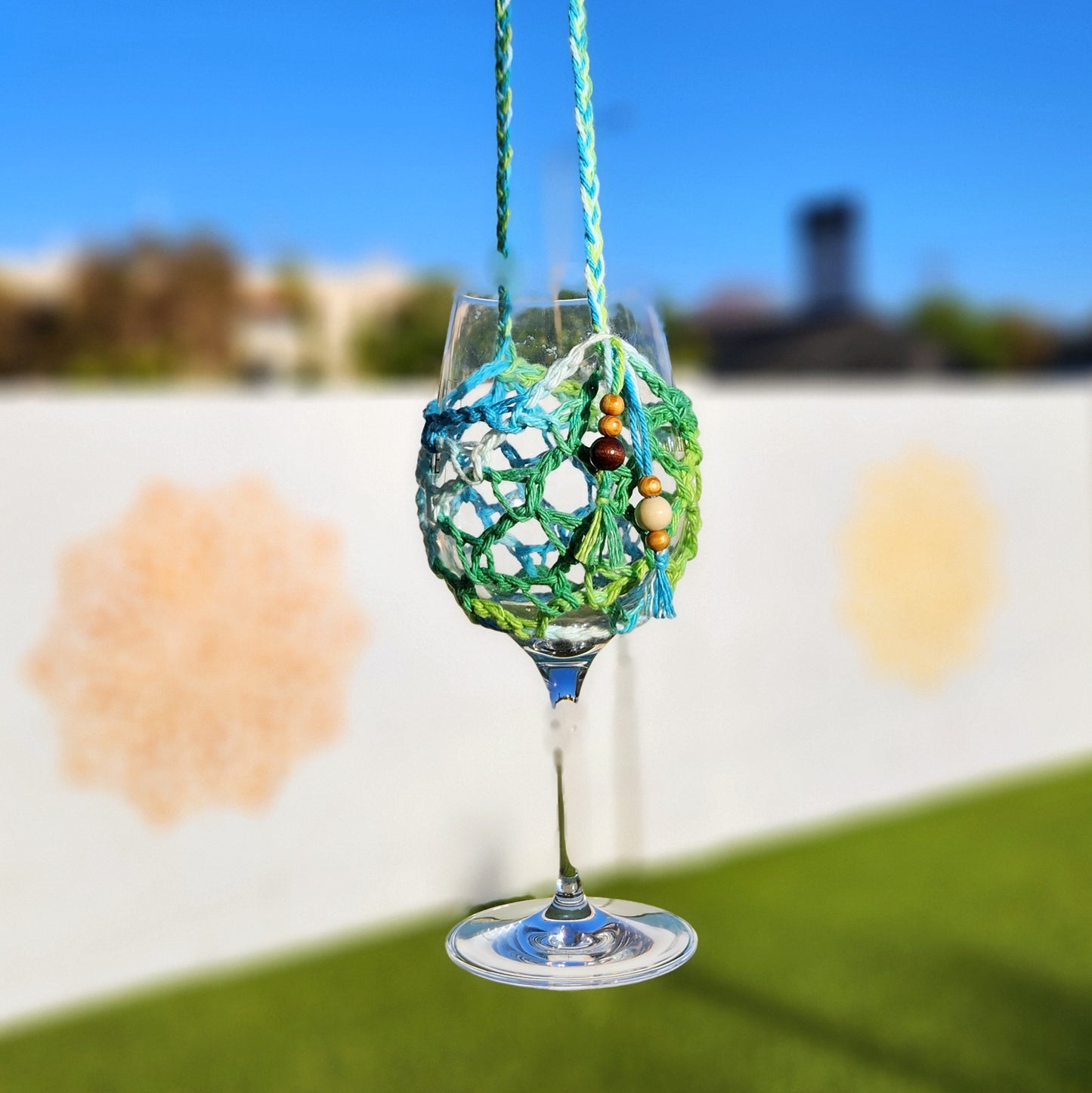 A wine glass suspended in a vibrant green and blue crocheted holder with a braided neck strap and wooden bead embellishments, against a bright blue sky.