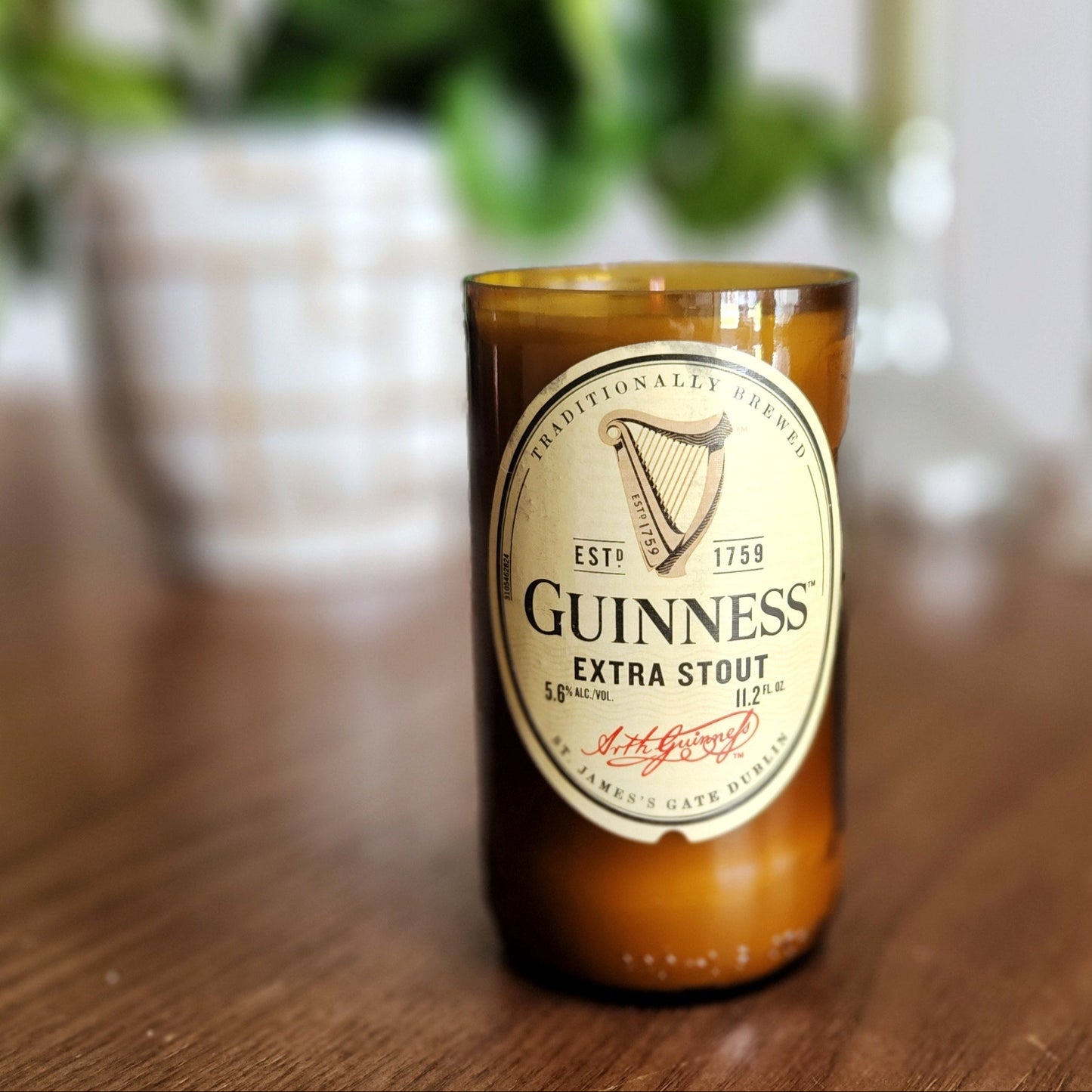 A hand-poured coconut soy wax candle crafted from a recycled Guinness Extra Stout beer bottle. The classic beige and black label features the iconic Guinness harp, adding a timeless Irish touch. This eco-friendly wooden wick candle has a polished glass rim, highlighting expert craftsmanship. Placed on a wooden surface with a softly blurred background of greenery and elegant candlesticks, this upcycled beer bottle candle is perfect for Guinness fans, craft beer lovers, and sustainable shoppers.